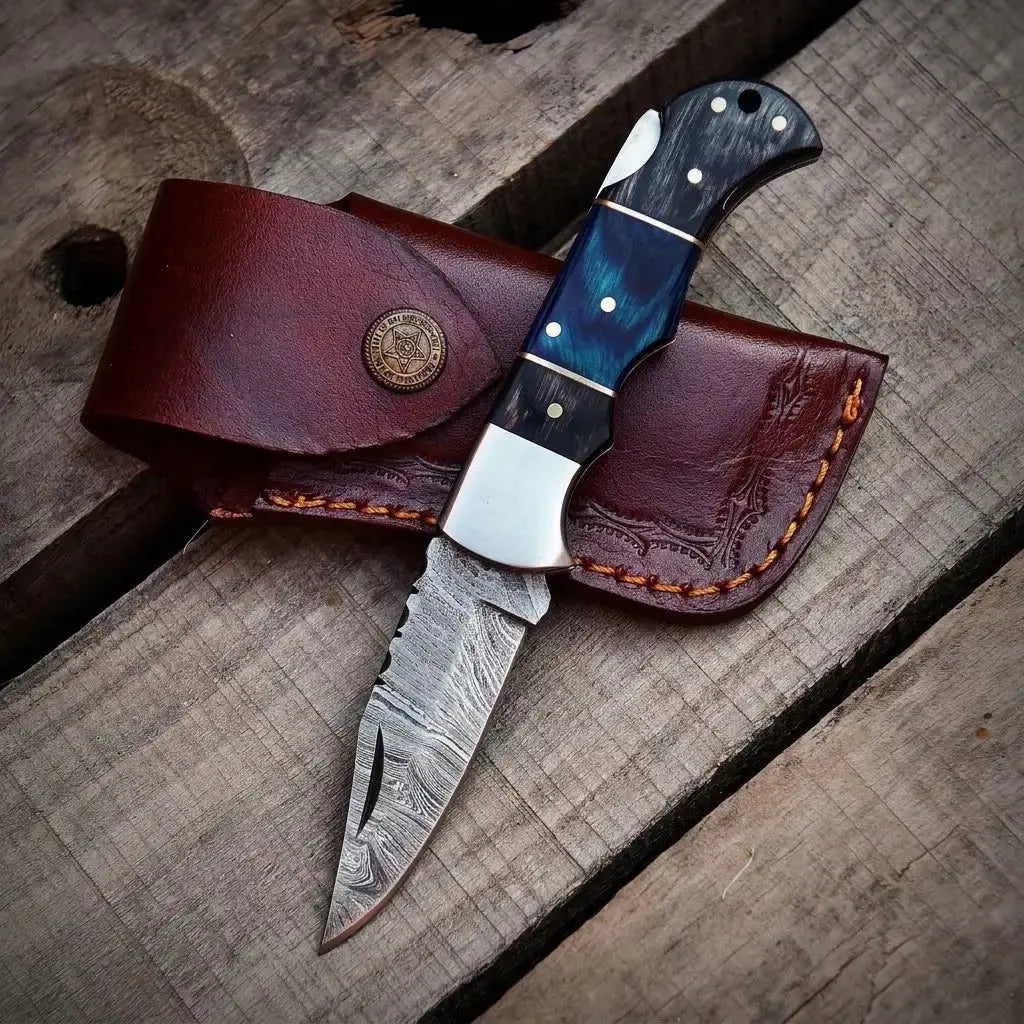 Handmade Damascus steel pocket knife with blue resin handle and custom brown leather sheath on wood background.